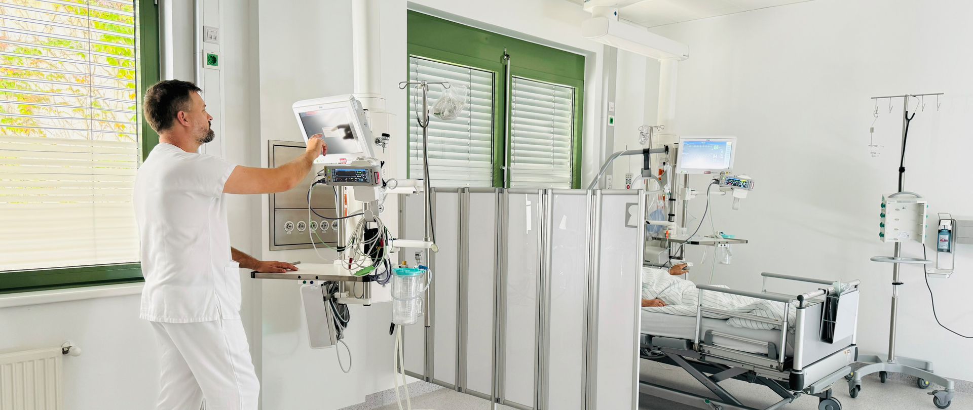 Ein Mann in weißer Kleidung steht an einem Monitor auf der Intensivstation, im Hintergrund ein Bett mit halb sichtbarem Patienten