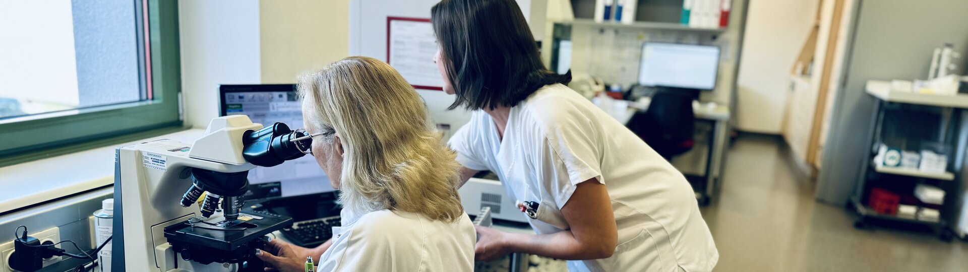 Eine Frau schaut in ein Laborgerät, die zweite steht daneben, beide Frauen sind von der Kamera wegschauend