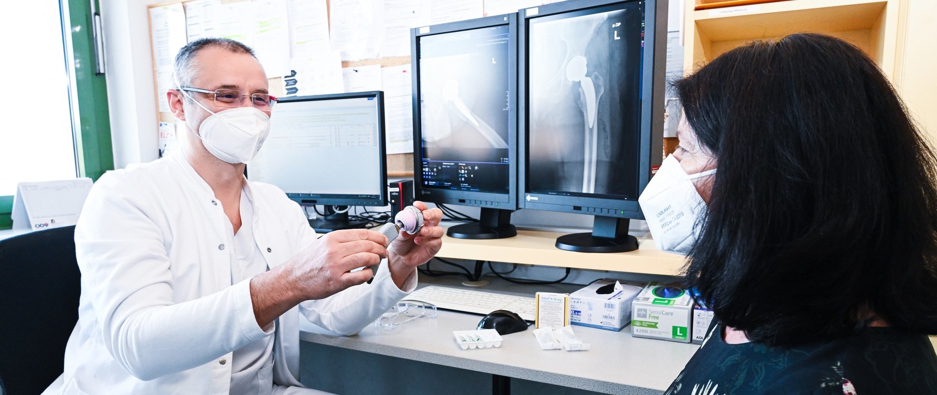 Ein Arzt mit Maske zeigt einer Frau mit Maske ein orthopädisches Gelenk